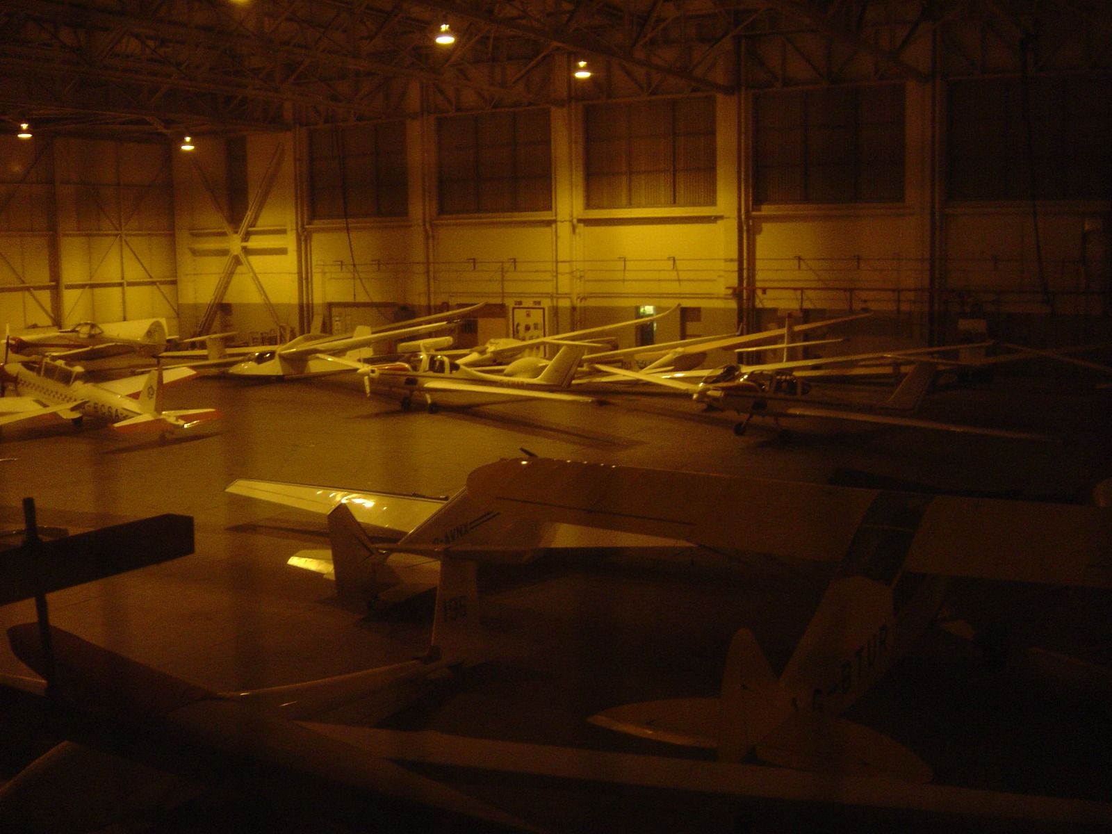 Rows of light aircraft and a glider parked in a dimly lit, cavernous historic hangar at night