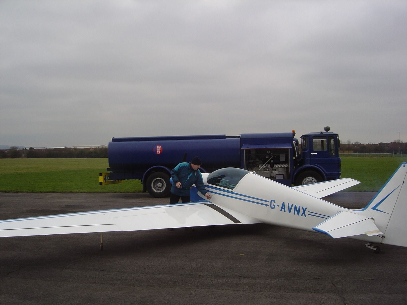 G-AVNX being refuelled from a large Avgas tanker truck on a grass airfield under grey skies
