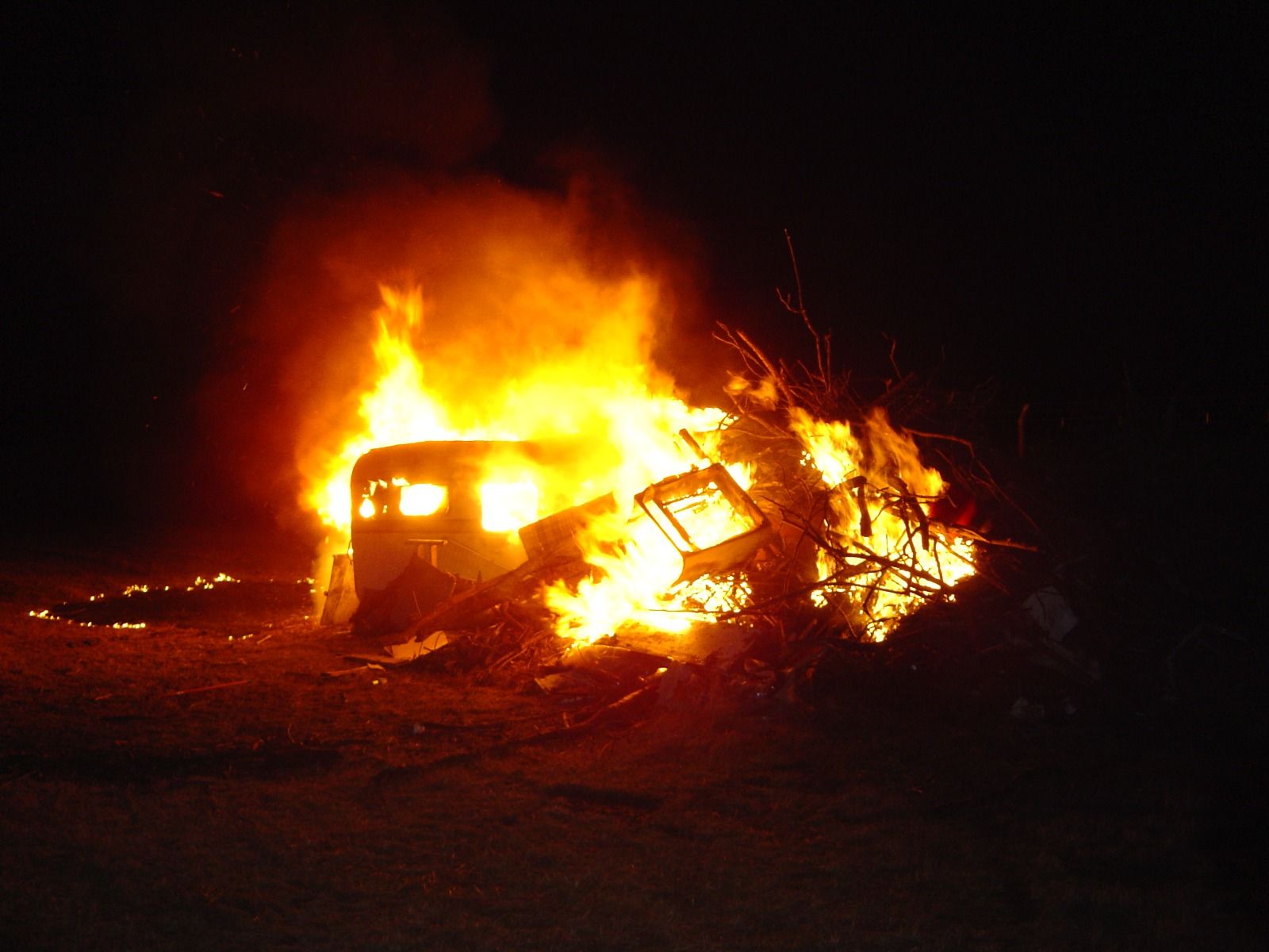 A large bonfire at night, consuming an old caravan and a pile of branches