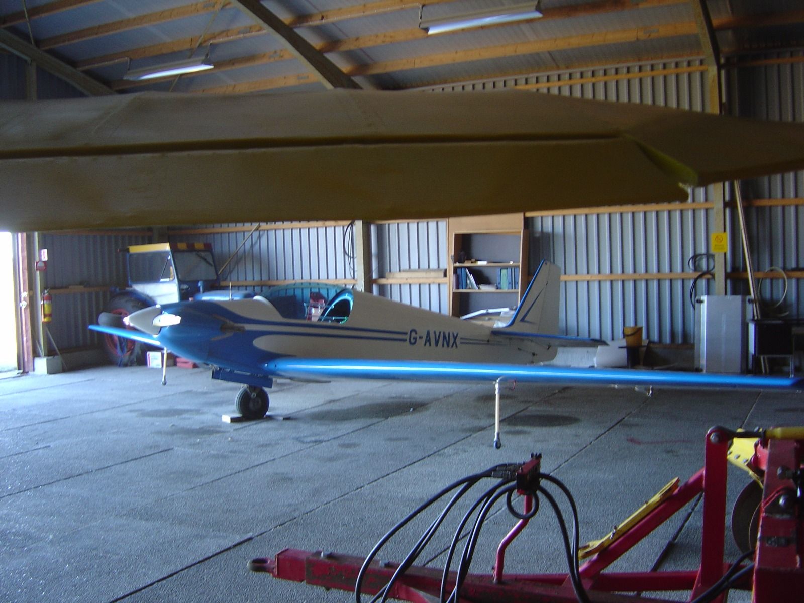 G-AVNX parked inside a modern farm-style hangar, viewed under the wing, with a tractor and shelving visible behind