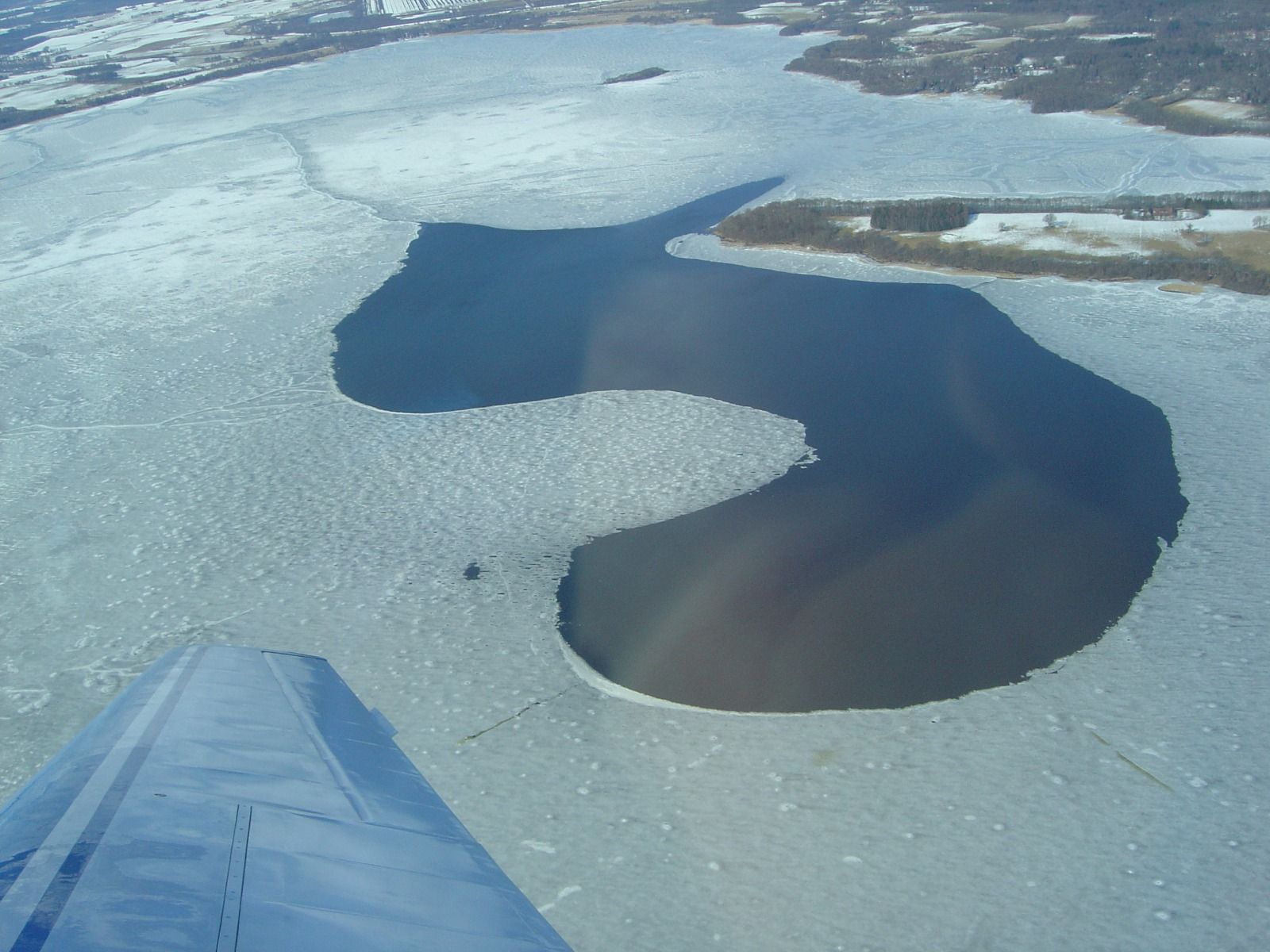 An aerial view of a partly frozen lake, with a dark patch of open water amid white ice, seen past the aircraft's wing