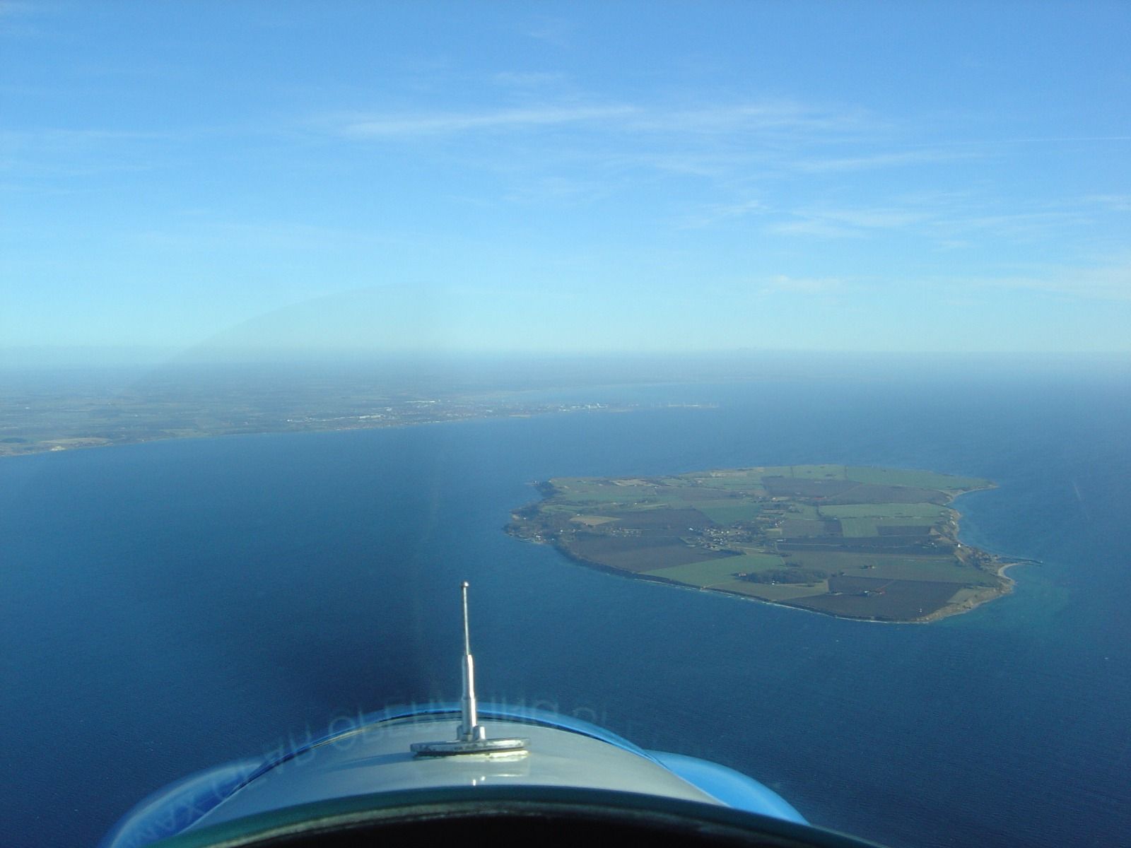 A clear blue-sky aerial view of a small green island surrounded by sea, seen from the cockpit of the aircraft