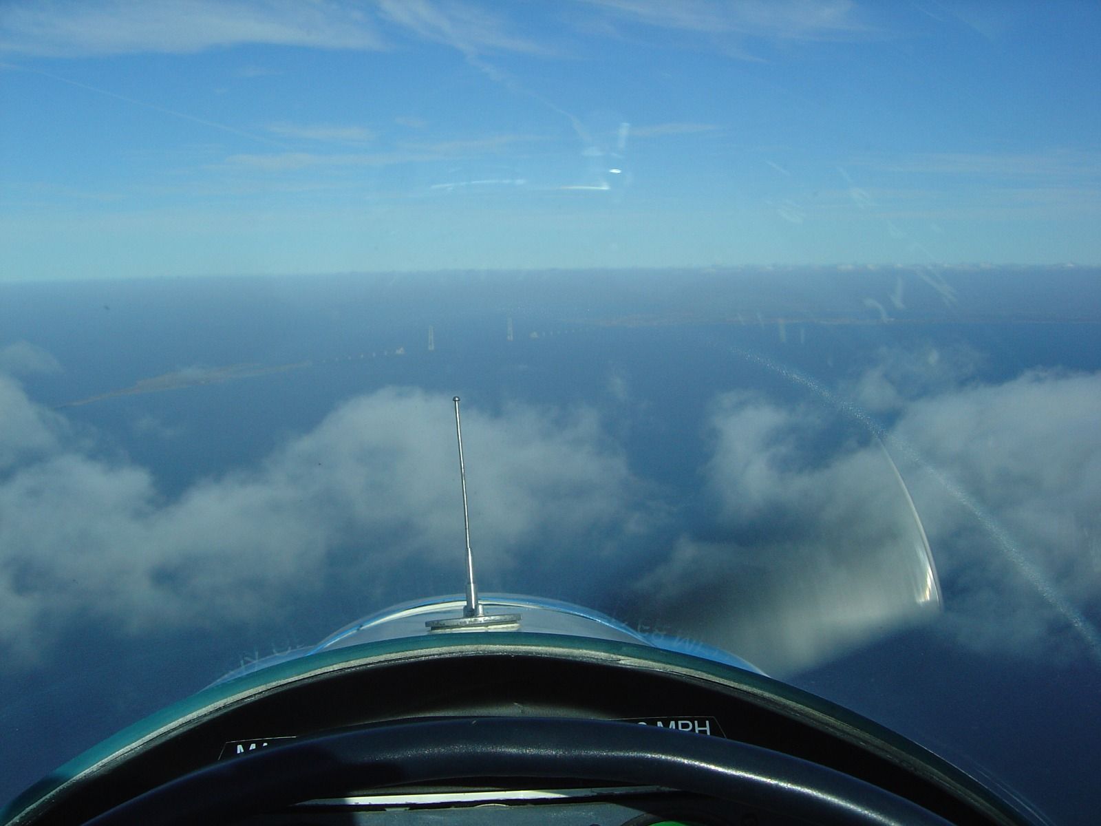 A hazy view from the cockpit over cloud toward the distant towers of a suspension bridge across water