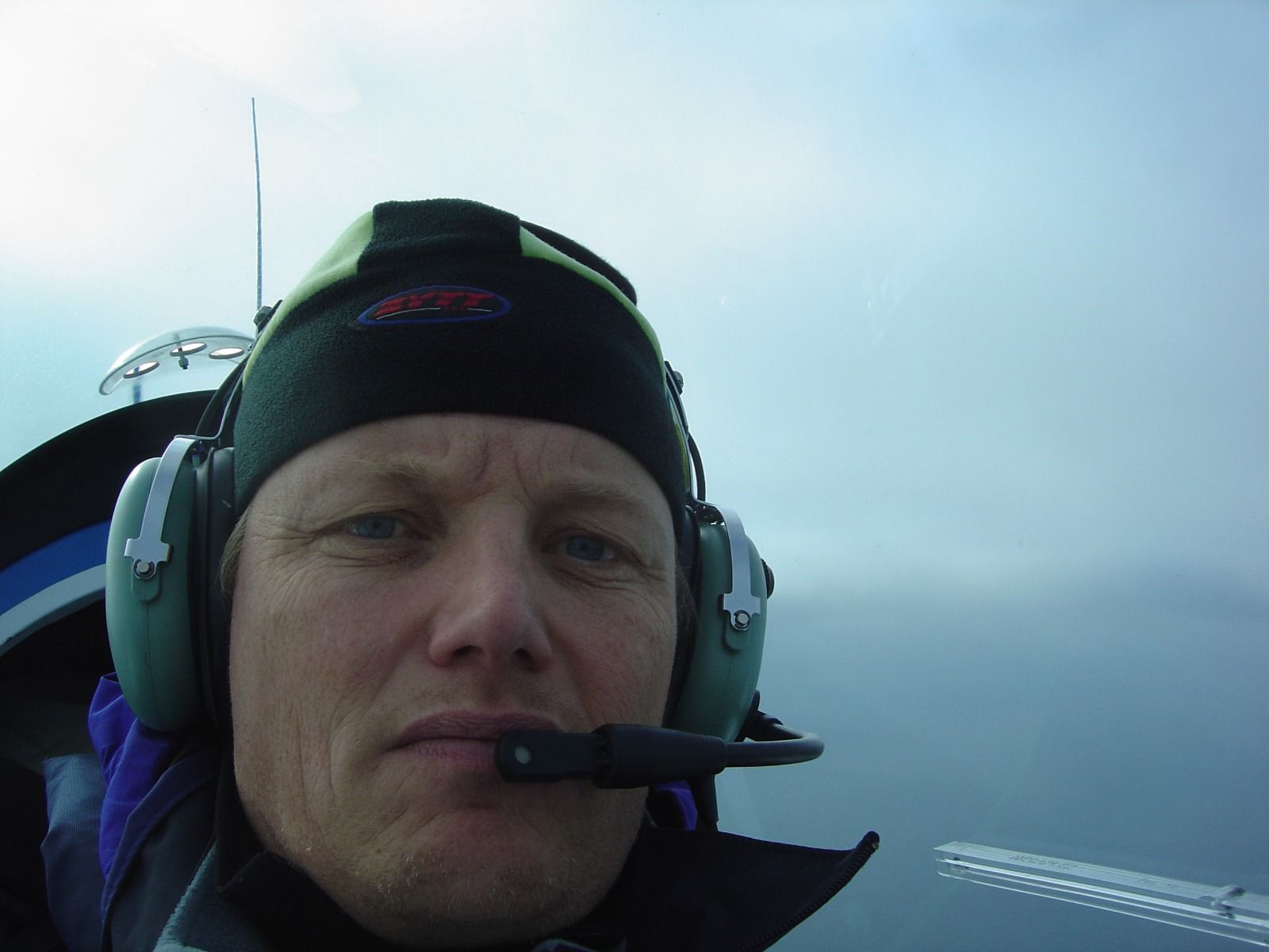 A pilot in a headset and beanie hat looking at the camera, with hazy sky visible through the canopy