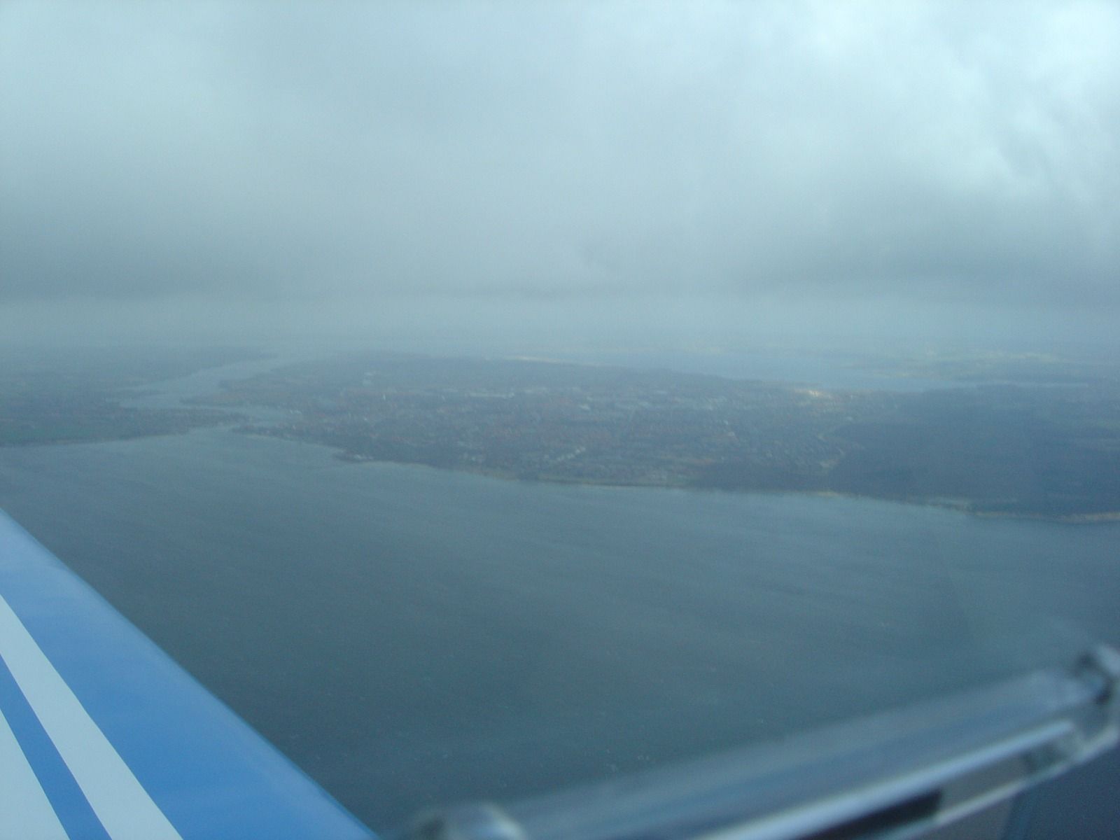 A hazy aerial view over water toward a low-lying coastline and built-up area under thick overcast