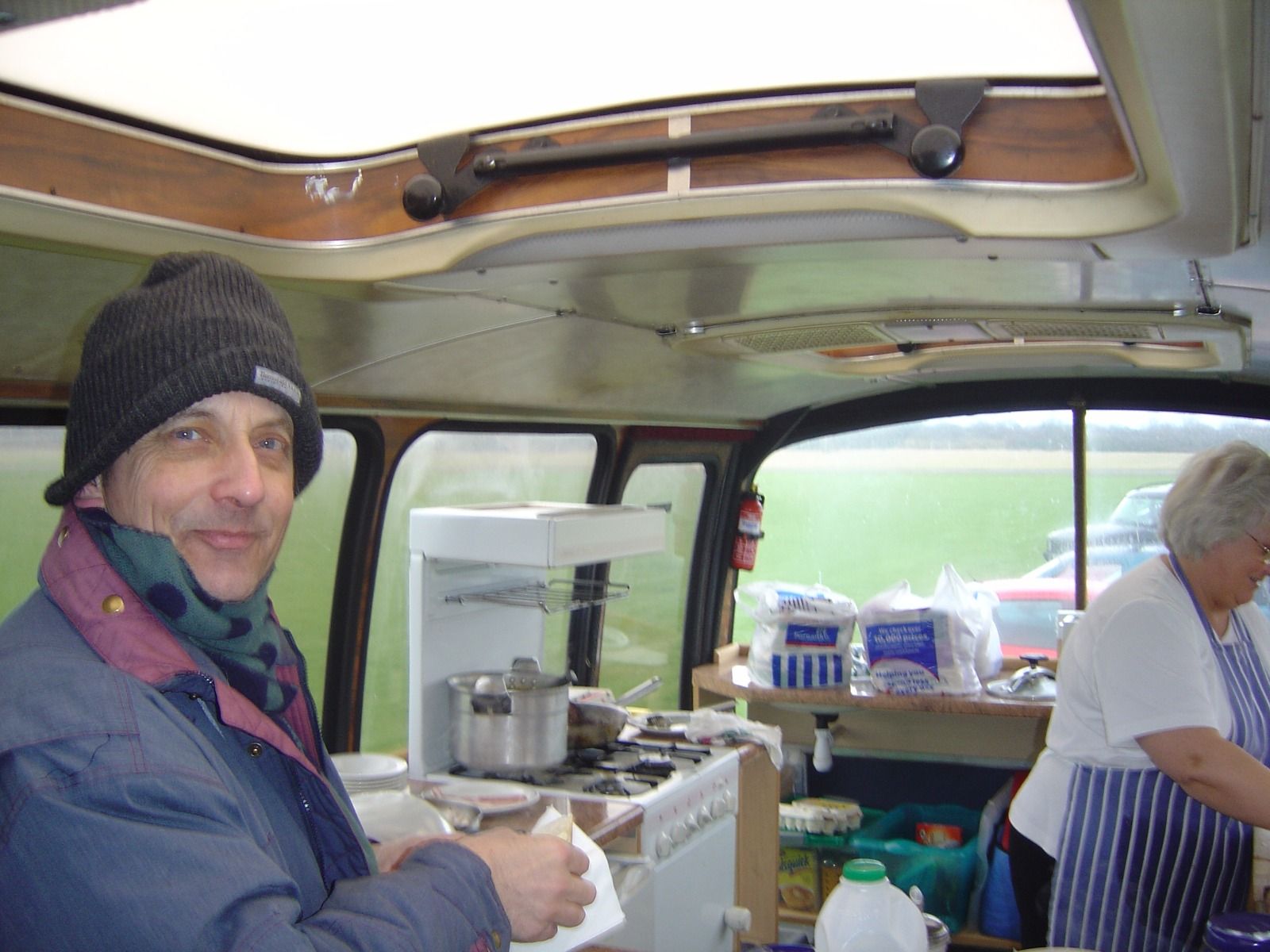 A man in a beanie and scarf inside a converted bus canteen, with a gas stove, pans and bread on the counter, and a lady in an apron behind him