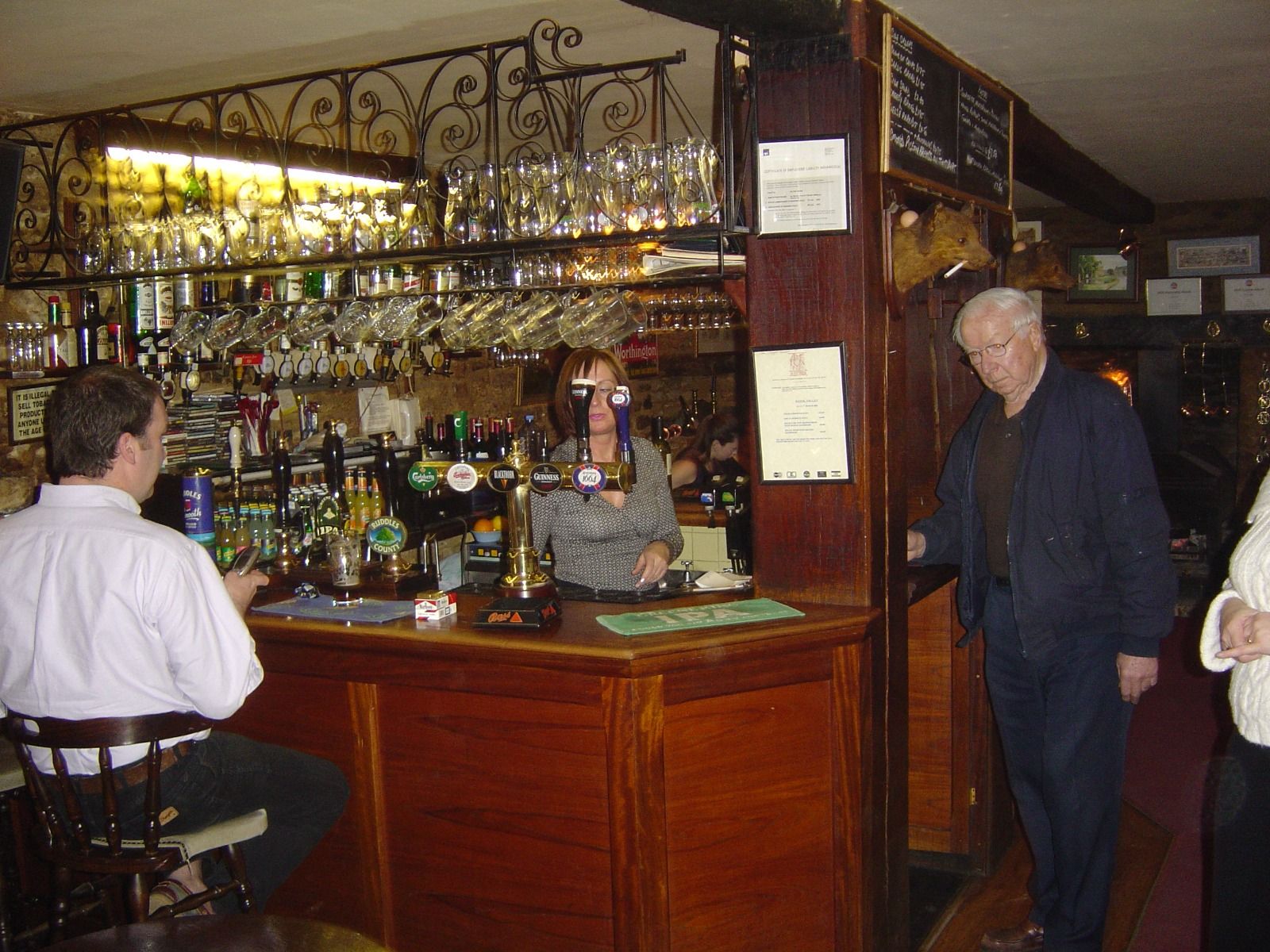 Two men at a traditional English pub bar with a barmaid pulling a pint, taps for Guinness and other beers, and a stuffed boar's head mounted on the wall