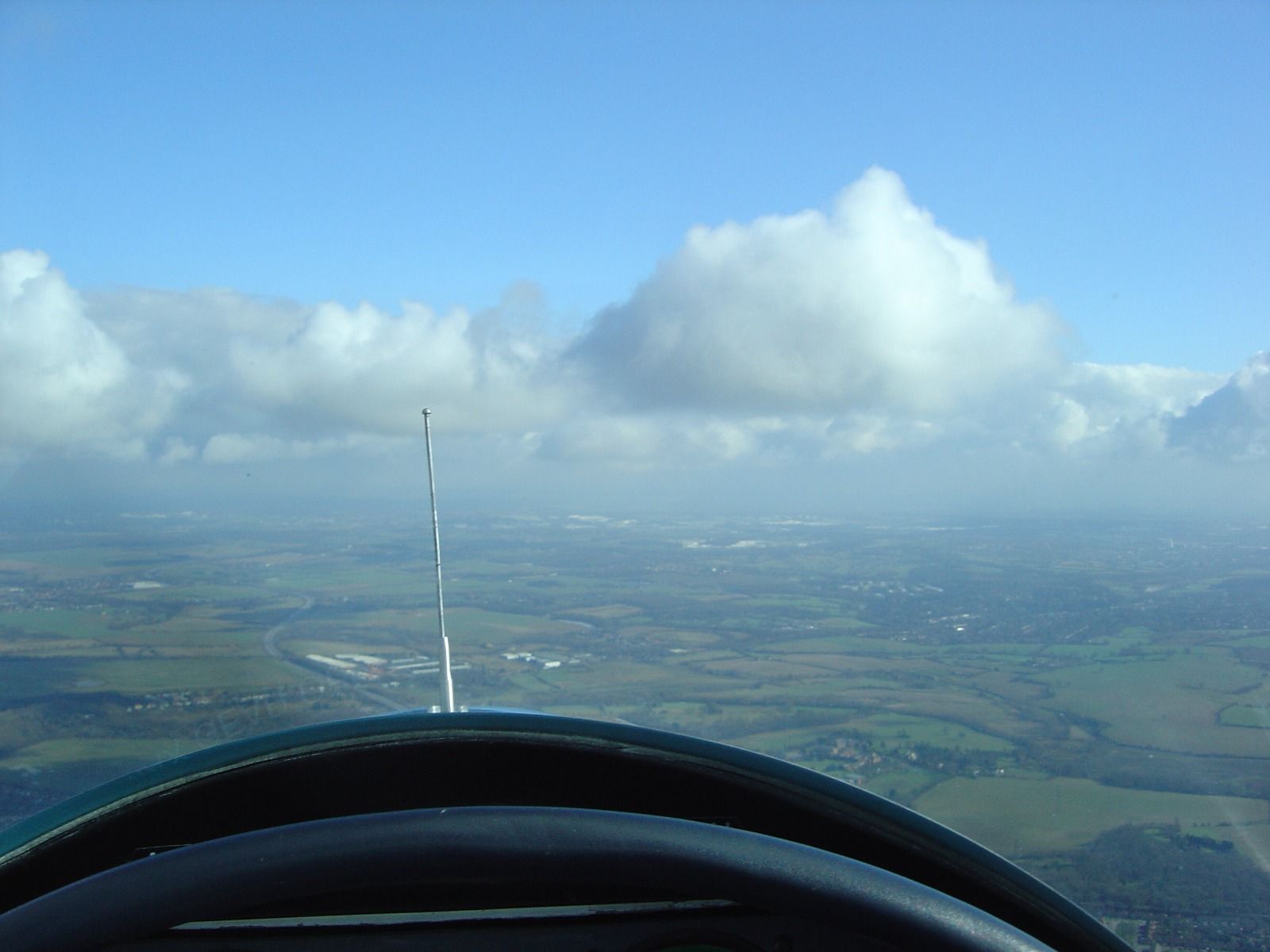 View from a cockpit of large, bright cumulus clouds building against a blue sky, over a hazy patchwork of fields