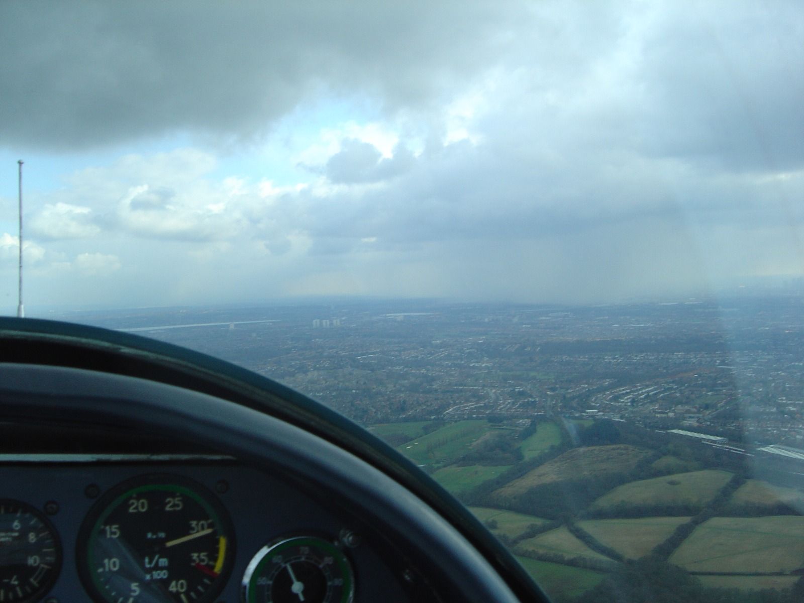 Cockpit view with an RPM gauge in the foreground and heavy dark rain clouds hanging over a hazy city in the distance