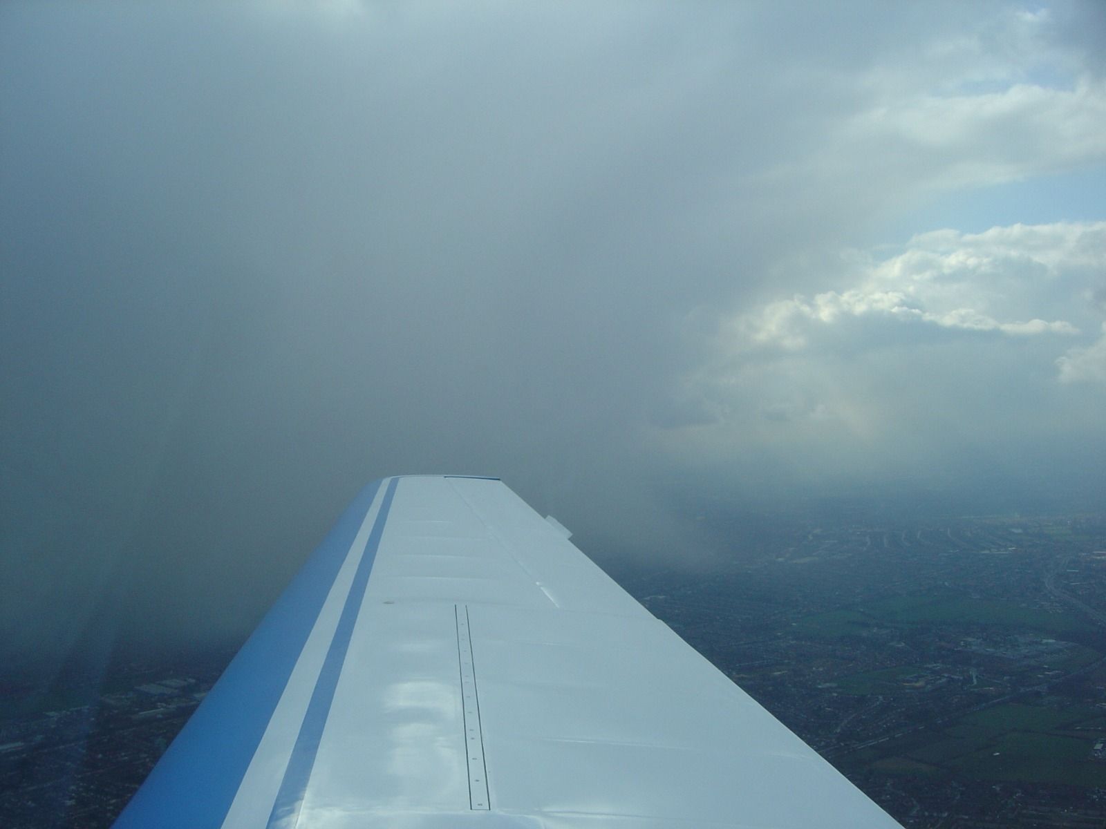 Aircraft wing with a dark rain cloud to the left and a gap of blue sky and lighter cloud to the right, over a sprawling city below