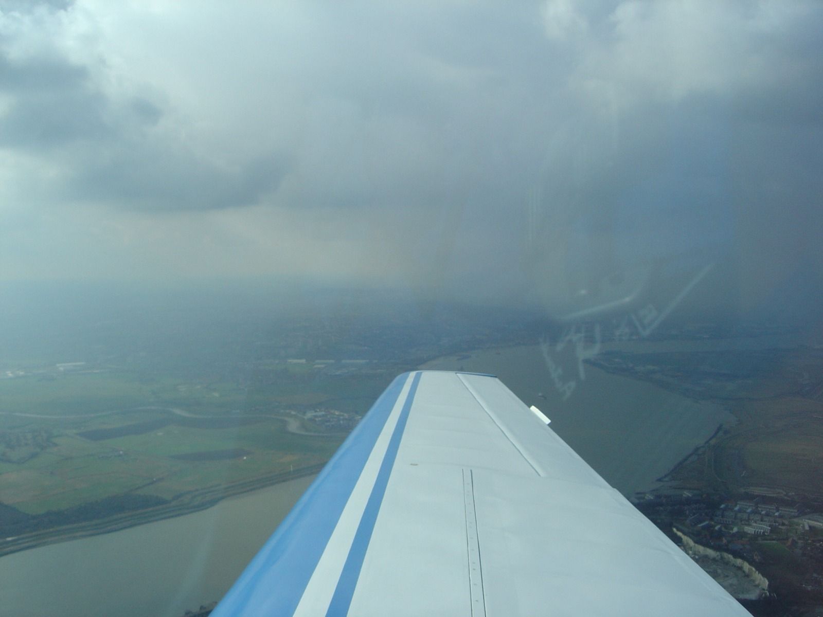Aerial view of a wide, muddy-brown river estuary with a hazy town on its far bank, seen past the aircraft's wing