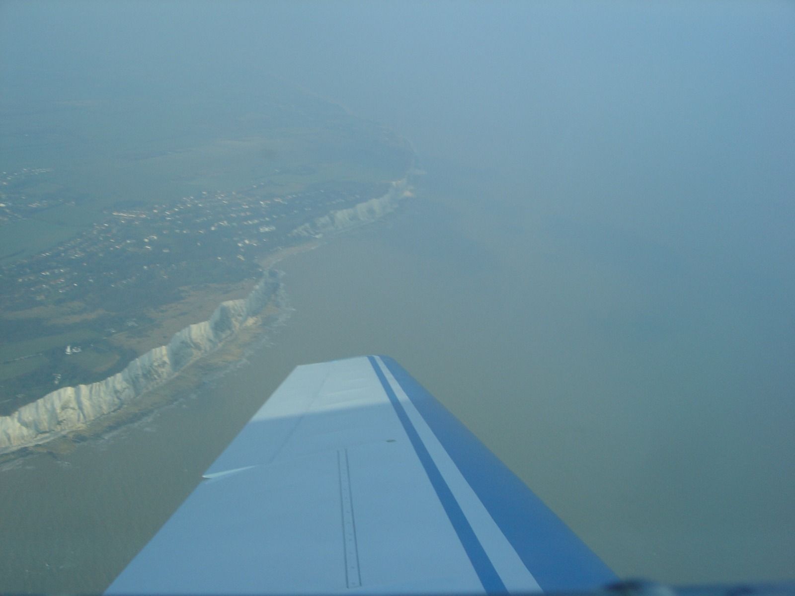 A clear aerial view of the White Cliffs of Dover from an aircraft wing, with a town and the sea beyond