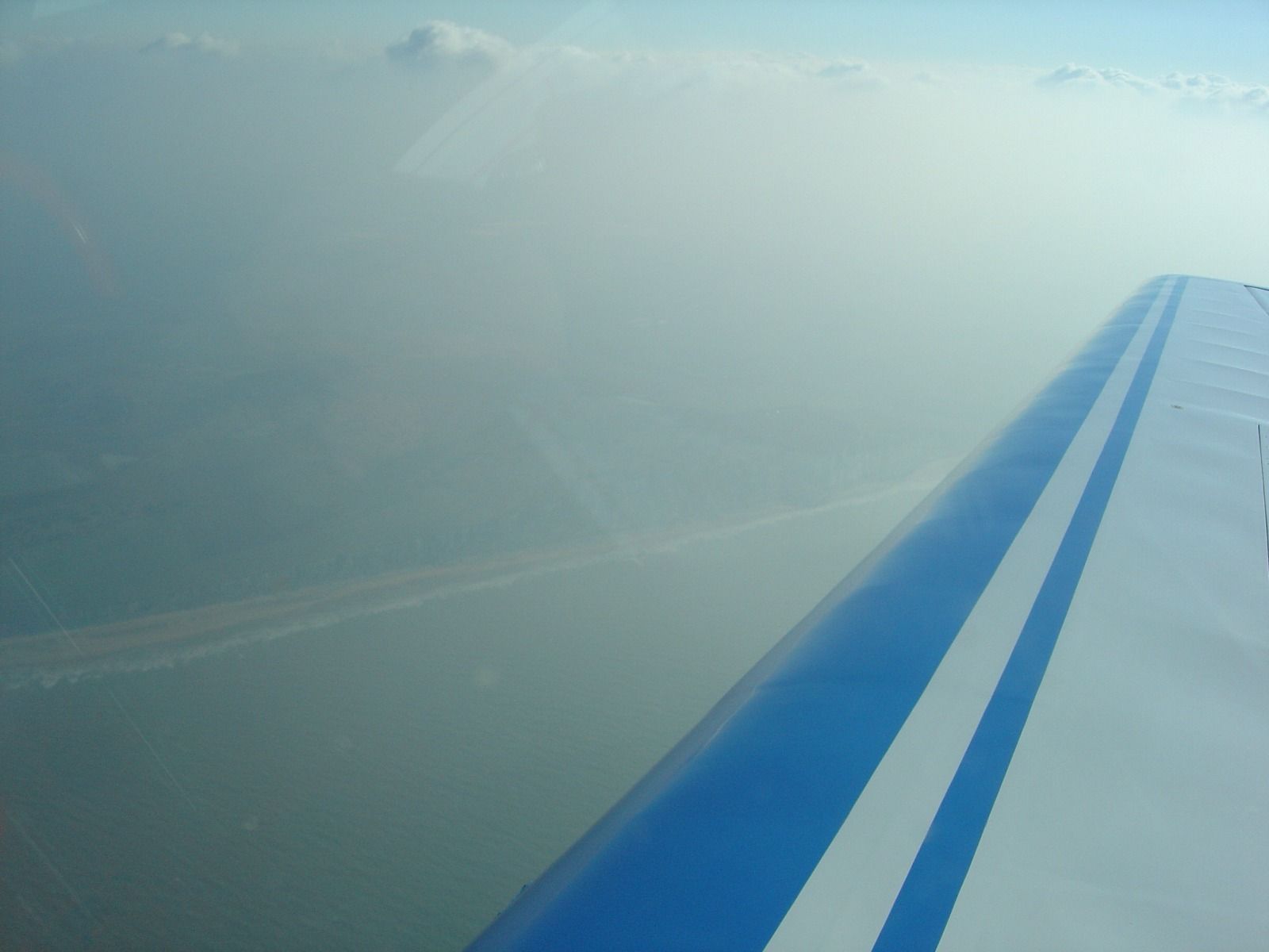 A hazy aerial view of a coastline with cliffs and a beach, seen from a wing under a partly cloudy sky