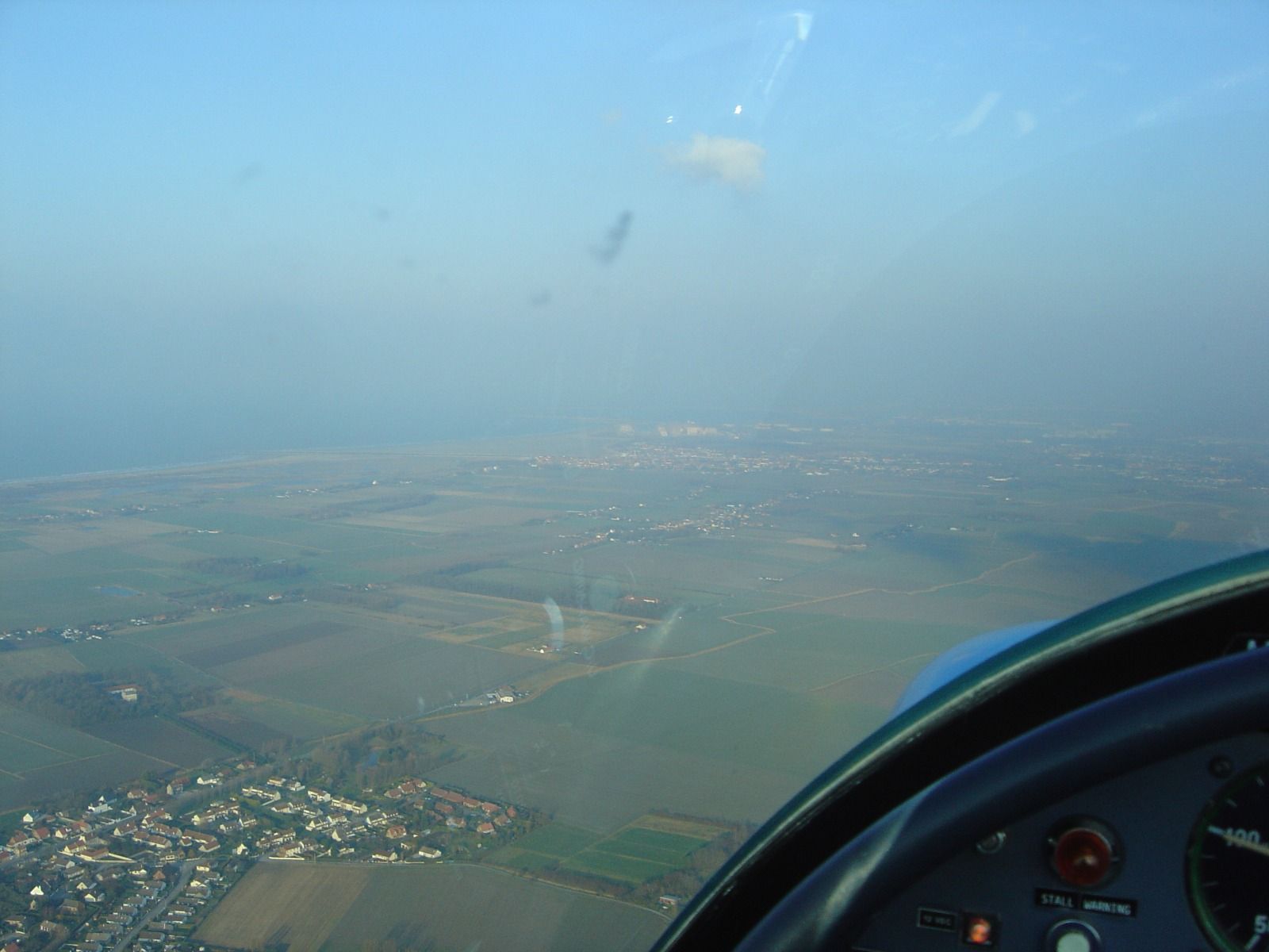 Cockpit view with a STALL WARNING placard on the panel, looking out over a hazy coastal town approaching from the sea