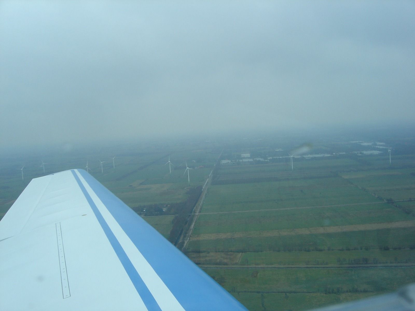 A wing view over flat, hazy farmland dotted with many wind turbines and a straight road running through it