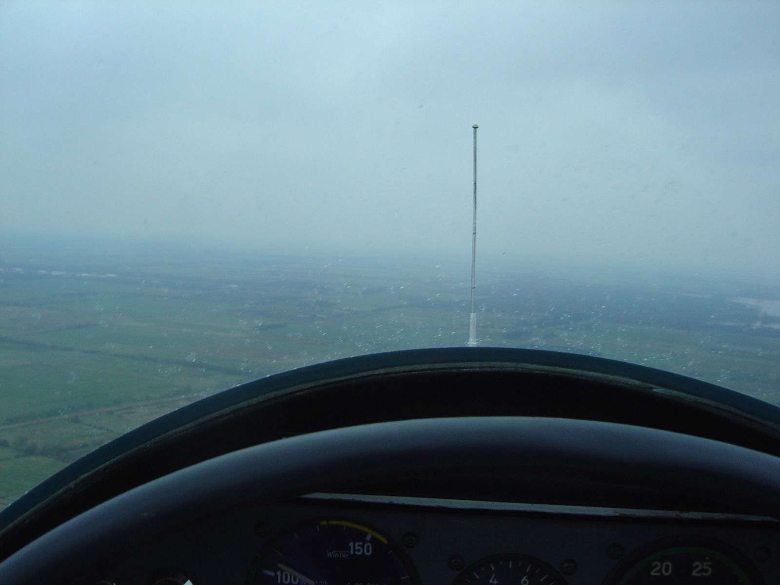 Cockpit view through a rain-speckled windscreen, with an airspeed indicator reading around 150 and hazy farmland below