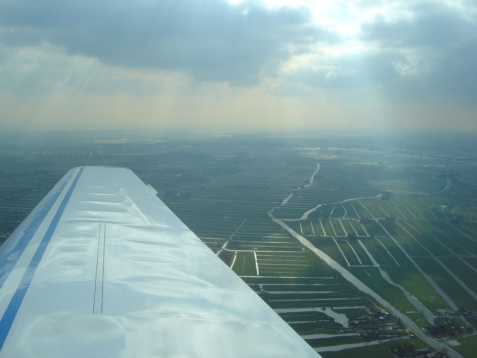 A wing view over a flat Dutch polder landscape crossed with dozens of drainage canals, with sunbeams breaking through clouds above