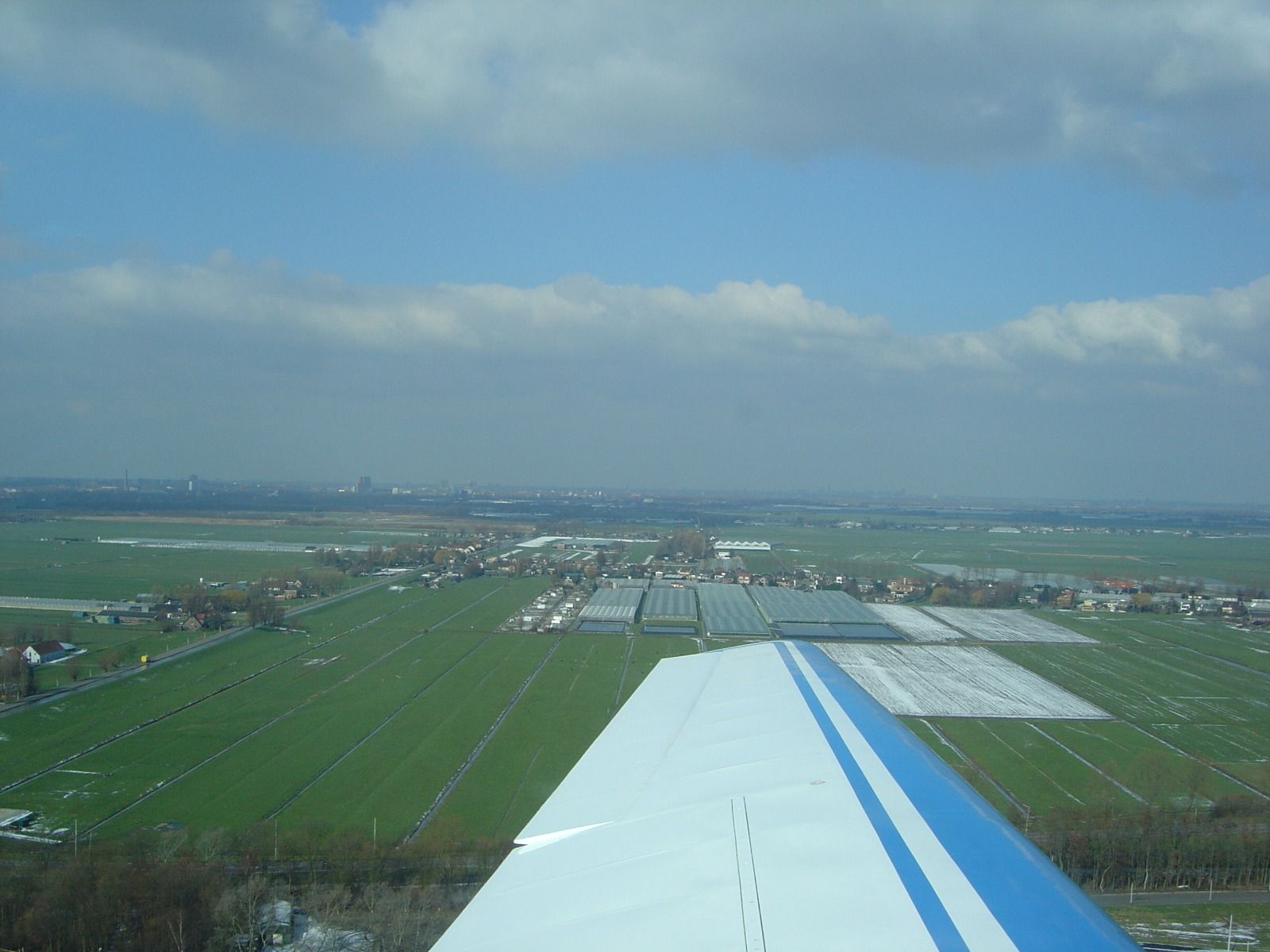 A wing view over green fields with patches of leftover snow, and a hazy city skyline in the far distance