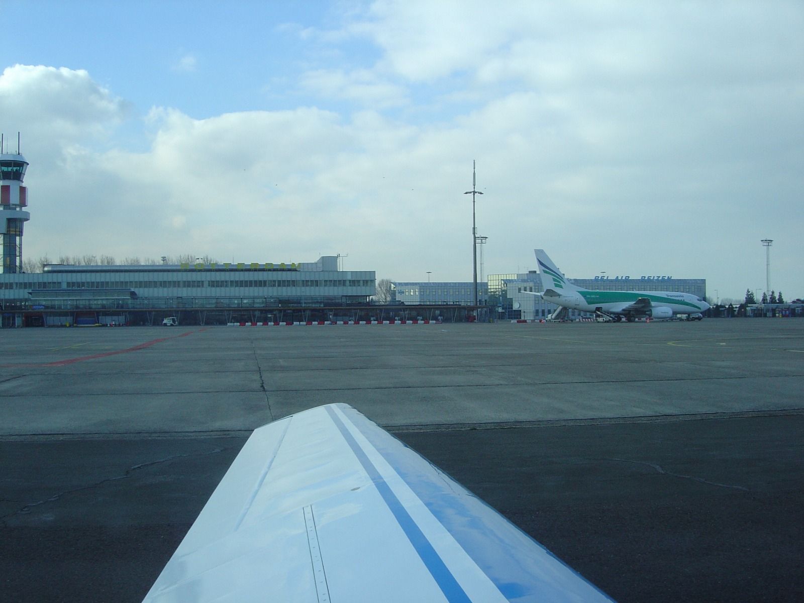 A wingtip in the foreground, with the Rotterdam Airport terminal building, control tower, and a green and white airliner on the apron behind