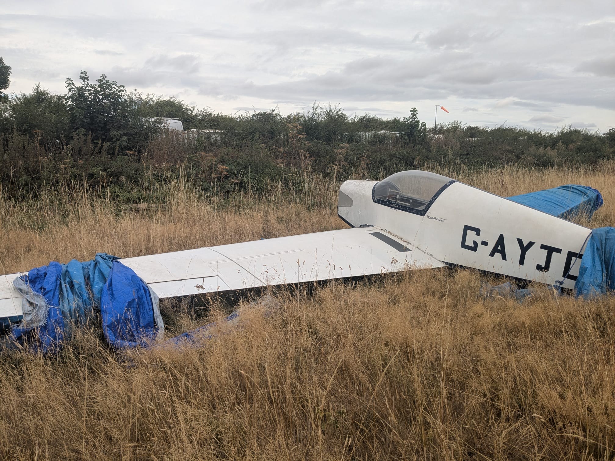 G-AYJD derelict in long grass, tarped over, windsock visible in the background