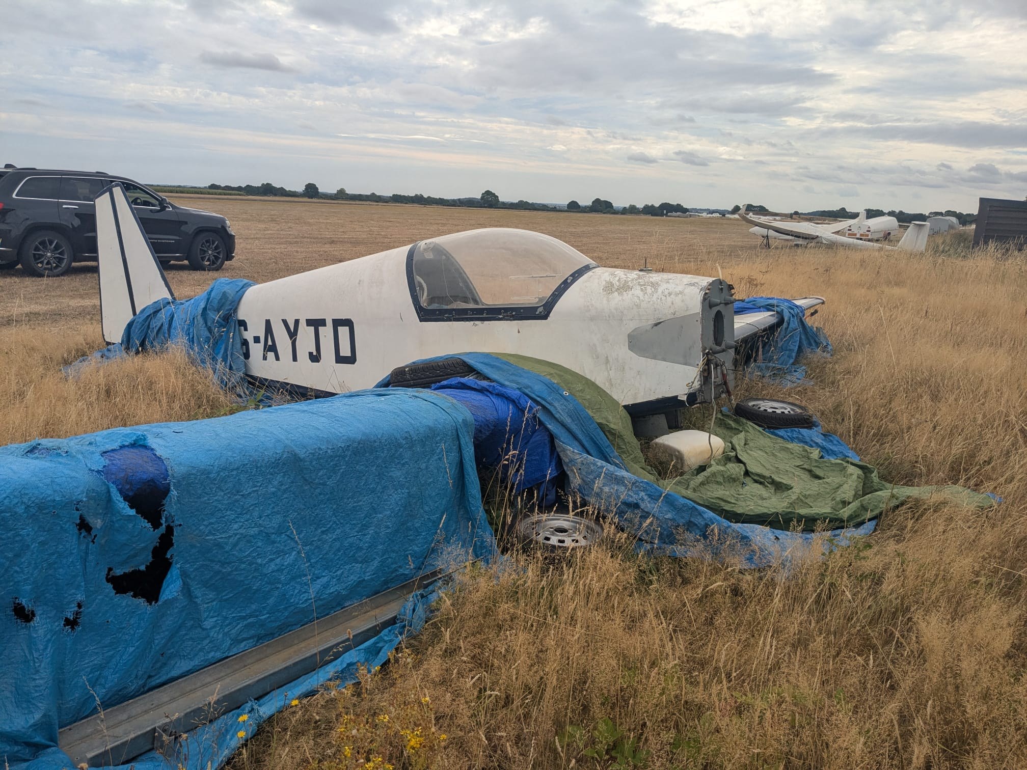 G-AYJD derelict in long grass, a car parked alongside and other gliders visible on the airfield behind